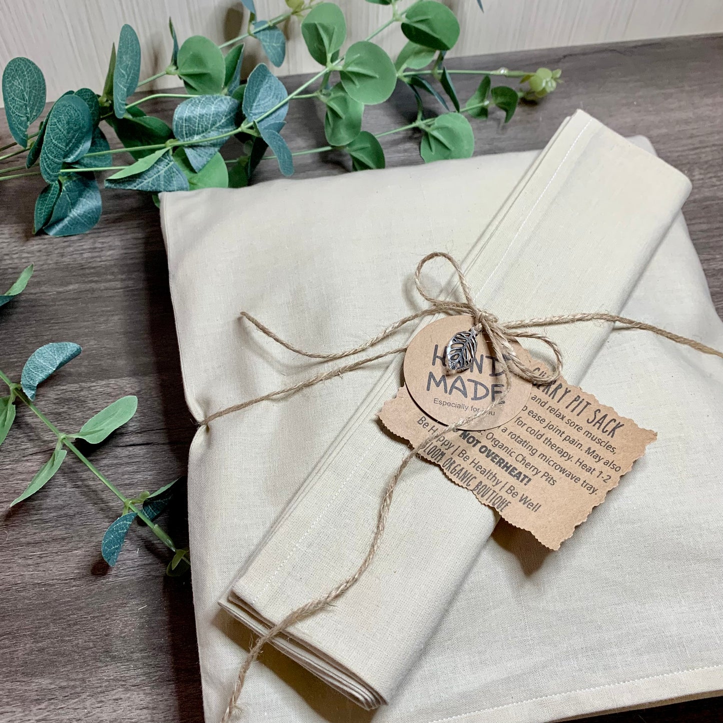 Wooden background with green eucalyptus leaves featuring an organic cherry pit sack. Made with organic cherry pits put into a case that is sewn in four sections for even heat distribution. Each sack comes with a case to protect the cherry pit sack.  