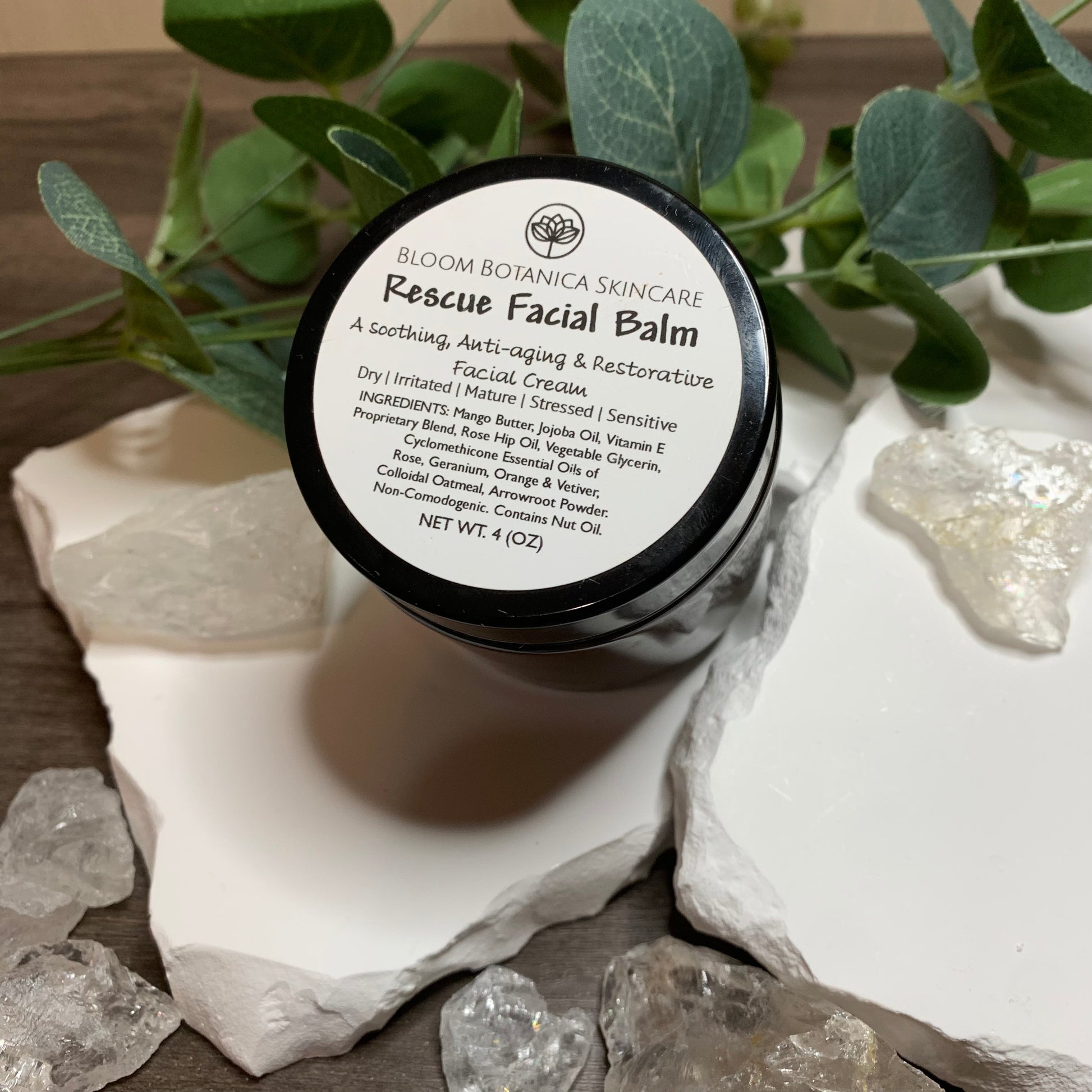 Rescue Facial Balm displayed with crystals and eucalyptus leaves. This balm is made from a base of mango butter, jojoba, and a vitamin E proprietary blend of skin loving goodness. It has the gentle, delicate smell of rose & citrus. It's so hydrating and calming to the skin and helps calm redness. 