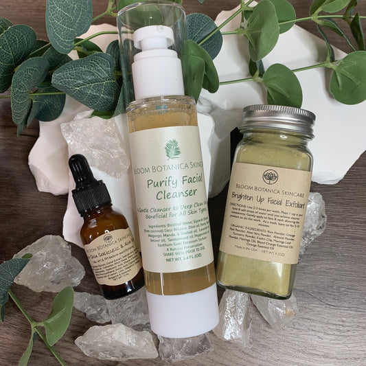 wooden background with greenery and three skincare products displayed. This is the Acne Skincare Bundle by Bloom Botanica Natural Skincare. his displays Hydrate Face Wash Brighten up exfoliant and Clear Skin Salicylic Serum.