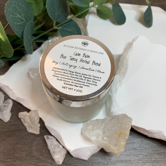 Beautiful wooden background with white stone, numerous crystal quartz and a eucalyptus branch all surrounding a 4 ounce jar of Calm Balm made with calming Blue Tansy. 