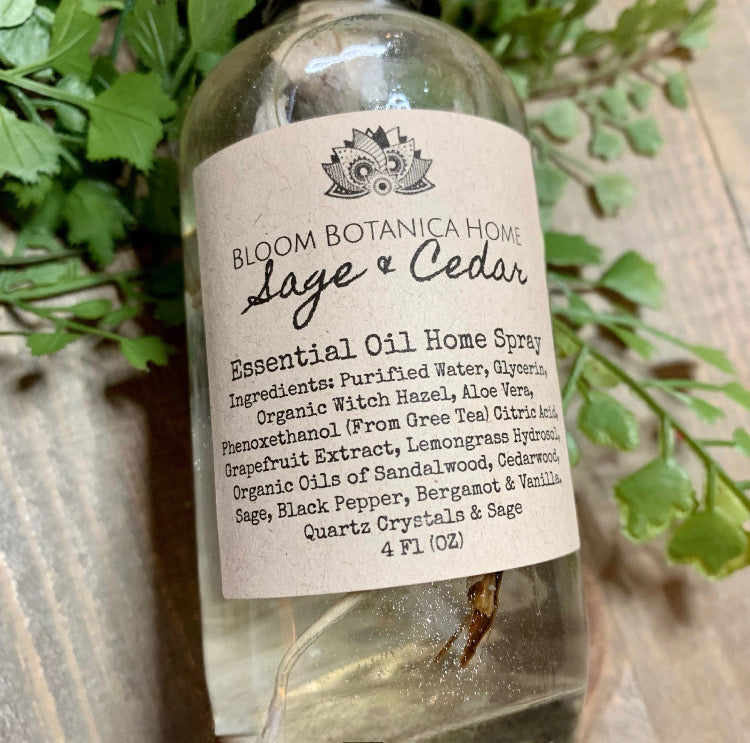 wooden background with green leaves featuring a beautiful bottle of Sage & Cedar Home fragrance spray. Comes with sage leaves and stem and crystals in the spray.