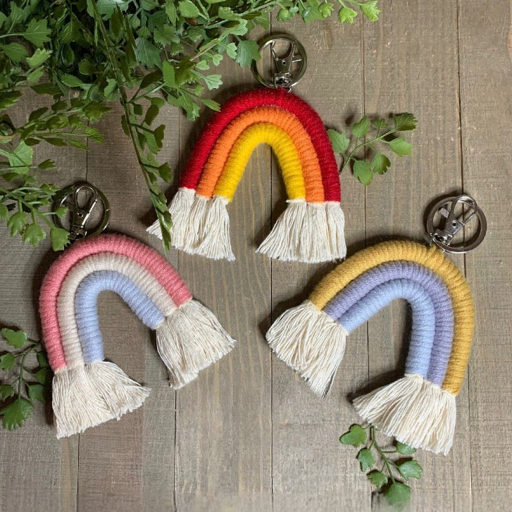 Cute macrame rainbow key chains, Purchase a set of three or one color. featured on a wooden background with green leaves. 