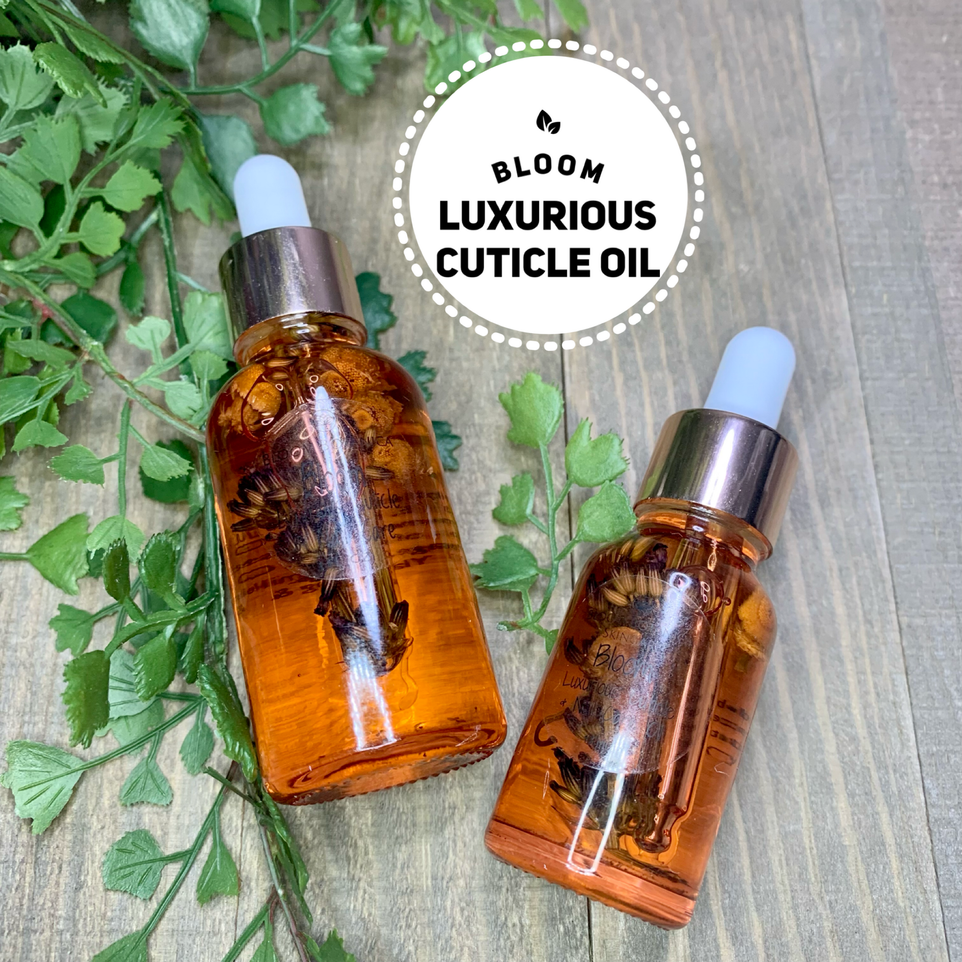 Wooden background with green leaves featuring two bottles of Bloom Cuticle oil. A 1 ounce and 15 ML size. This cuticle oil is so pretty. it contains lavender stems and chamomile buds. it looks like the flowers are blooming in the bottle.