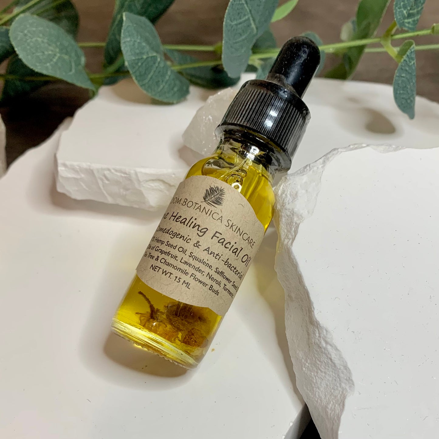 Acne healing Facial Oil is an anti-bacterial gentle blend of essential oils and herbal extracts that ward off acne and promote healing to the skin. It is displayed on white stone with a wooden background, crystals and eucalyptus leaves